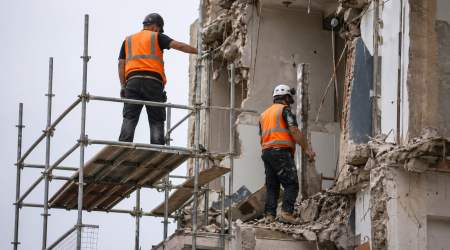Two construction workers wearing personal protective equipment, including hard hats and hi-vis vests, working safely on scaffolding at a demolition site.
