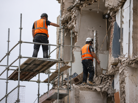 Two construction workers wearing personal protective equipment, including hard hats and hi-vis vests, working safely on scaffolding at a demolition site.