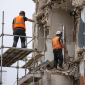 Two construction workers wearing personal protective equipment, including hard hats and hi-vis vests, working safely on scaffolding at a demolition site.
