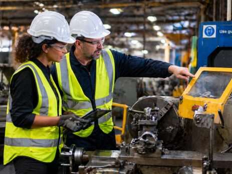 Two factory workers in protective gear discussing safety features on a metal lathe, highlighting machine guarding and operational safety.