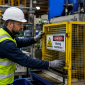 A male factory worker in a hard hat and hi-vis vest carefully checks the safety guard on a piece of industrial machinery on a busy factory floor.