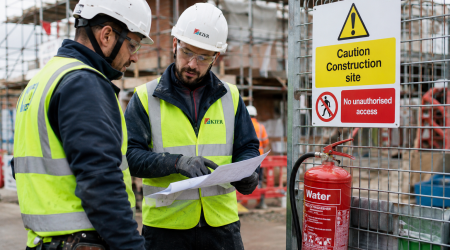 Two construction workers wearing personal protective equipment, discussing safety procedures on a busy UK construction site, with safety equipment visible.