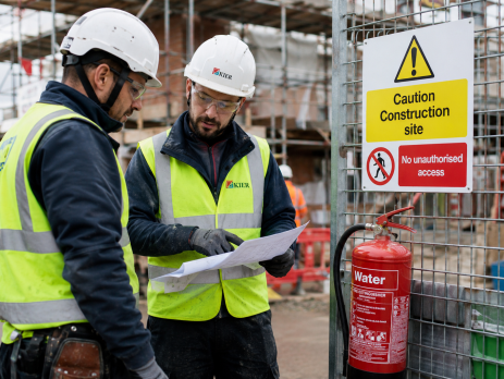 Two construction workers wearing personal protective equipment, discussing safety procedures on a busy UK construction site, with safety equipment visible.