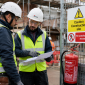 Two construction workers wearing personal protective equipment, discussing safety procedures on a busy UK construction site, with safety equipment visible.